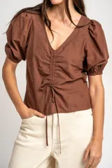 Blusa marrón de algodón con bordados sutiles, escote en V, mangas cortas abullonadas y detalle de fruncido con cordón ajustable en el centro del frente.