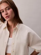 Camisa de denim de corte cropped y mangas cortas, con cuello clásico y cierre frontal de botones. Presenta un diseño minimalista y estructurado.