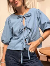 Blusa de lino color celeste con mangas abullonadas tres cuartos y cierre frontal mediante tres lazos decorativos tipo moño.