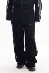Pantalón deportivo negro de calce relajado con cintura elástica. Presenta una franja lateral gruesa de color gris claro con bordes blancos. Se observa un pequeño bordado en la zona del bolsillo con la palabra "Delosalto".