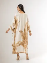 Vestido maxi túnica de corte oversized, color beige y blanco con estampa de hojas de palmera en tonos camel, mangas murciélago y bolsillos laterales.