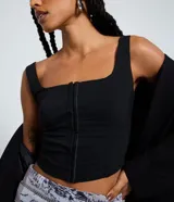 Top negro ajustado al cuerpo con escote cuadrado y breteles anchos. Presenta un cierre frontal con colchetes visibles.