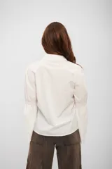 Camisa blanca de corte clásico con cuello camisero y cierre frontal de botones. Destaca por sus mangas largas con detalle de frunce a la altura del codo y terminación acampanada con bordado inglés.