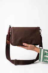 Cartera bandolera Rotunda modelo Naru, de color marrón, con solapa frontal y tirante ajustable. Incluye detalle colgante de cordones trenzados con cuentas decorativas y el logo de la marca en letras metálicas.