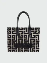 Tote bag Balmain de tamaño pequeño con estampado de monograma geométrico en blanco y negro. Presenta asas de cuero negro, detalles de costuras en contraste en los laterales y una placa de cuero con el logo de la marca grabado en la parte frontal.