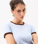 Remera tipo baby tee de corte ajustado y manga corta, en color beige claro, con ribetes en tono marrón oscuro en el cuello redondo y los puños.