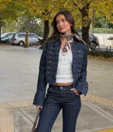 Chaqueta de denim estilo militar con diseño napoleónico, caracterizada por su doble fila de botones metálicos decorativos en el frente y mangas acampanadas con detalles de botones en los puños. Presenta un corte entallado y cuello redondo.