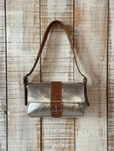 Cartera tipo bandolera modelo Baby Jade, confeccionada en material sintético plateado metalizado con detalles en cuero marrón texturado, incluyendo una tira central con hebilla metálica y correa de hombro ajustable.