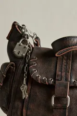 Cartera mini de cuero marrón con terminación gastada, cuerpo blando, bolsillos exteriores y solapa con cierre de hebilla. Incluye herrajes metálicos envejecidos y detalles colgantes con candado y llave.