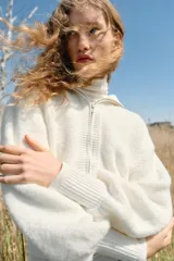 Chaqueta de punto color blanco con cuello subido, cierre frontal de cremallera metálica y mangas largas abullonadas con puños ajustados.