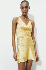 Vestido corto de satén con estampado de lunares, escote fluido tipo cowl neck y tirantes finos que se cruzan en la espalda con una aplicación metálica circular.