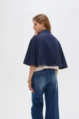 Poncho corto de denim azul con cuello alto, cierre frontal y diseño amplio de inspiración capa.