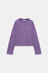 Camiseta de manga larga con corte oversize, confeccionada en algodón color violeta con efecto lavado. Presenta cuello redondo, bajo asimétrico y detalles de texto con aplicación de brillos en el pecho y las mangas.