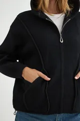 Abrigo negro con capucha, confeccionado en tejido de textura suave. Presenta cierre frontal con cremallera, bolsillos laterales integrados con costuras decorativas y puños ajustados.