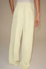 Pantalón de tiro medio de pierna ancha color amarillo pastel, confeccionado con mezcla de lino. Cuenta con bolsillos laterales ocultos, pliegues delanteros y cierre frontal con cremallera, botón y ganchos metálicos.