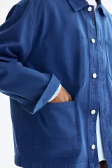 Sobrecamisa de lona de algodón en color azul, con cuello clásico, cierre frontal de botones y tres bolsillos de parche en la parte delantera. Presenta mangas largas con puños abotonados y canesú en la espalda.