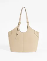 Cartera tipo shopper color beige, con diseño de costura central vertical y detalles de tachas metálicas doradas en los laterales superiores. Cuenta con correas ajustables mediante hebillas metálicas y logo grabado en la parte inferior.