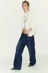 Blusa de bambula color blanco con cuello subido y manga larga. Presenta detalles de volantes y puntillas a tono en el frente, con cierre posterior mediante abertura y botón.