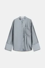 Camisa de cuello mao y manga larga con puños anchos, de color gris satinado. Cuenta con un bolsillo de plastrón en el pecho con detalles bordados a tono, al igual que los puños, y cierre frontal con botones ocultos bajo solapa.