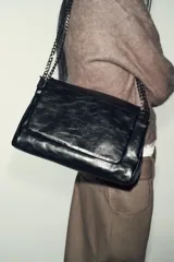 Bolso de hombro con solapa, confeccionado en material sintético con acabado brillante y efecto envejecido. Presenta un diseño estructurado con costuras decorativas, tres compartimentos internos, uno de ellos con cierre de cremallera, y asa de cadena metálica. Se cierra mediante un imán oculto.