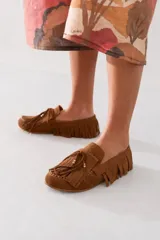 Mocasín de gamuza color marrón, con diseño de flecos decorativos en todo el contorno y la capellada. Presenta cordones decorativos con detalles metálicos dorados en las puntas.