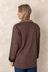 Blazer de gamuza color tostado con diseño artesanal. Presenta flecos en las solapas y bolsillos, bordados florales en los hombros con detalles de perlas colgantes y aplicaciones decorativas en los bolsillos.