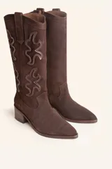 Bota de caña alta estilo tejano confeccionada en gamuza color marrón oscuro. Presenta detalles de costuras decorativas en forma de llama en la caña, taco cuadrado de madera y punta almendrada.
