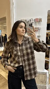 Camisa de corte relajado confeccionada en algodón con estampado de cuadros tartán en tonos marrones y beige. Presenta cuello clásico, cierre frontal de botones, mangas largas con puños abotonados y un bolsillo frontal en el pecho.