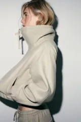 Sudadera tipo bomber de tejido suave, color beige, con cuello alto ajustable mediante cordones, cierre frontal con cremallera, mangas largas con puños elásticos y bolsillos laterales de vivo.