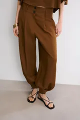 Pantalón de tiro alto en color beige, con diseño de pierna holgada y puños elásticos con cierre de botón en el bajo. Presenta cierre frontal con cremallera y doble botón en la cintura, además de bolsillos laterales.