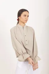 Blusa de diseño cuadrillé en tonos beige y verde claro, de corte amplio y mangas largas abullonadas con puños elastizados con volados. Cuenta con cuello mao y una abertura frontal parcial con botones.
