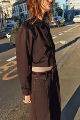 Chaqueta corta de color marrón oscuro, con cuello de solapa, manga larga con trabillas en los puños, bolsillos delanteros con solapa y cierre frontal con botones.