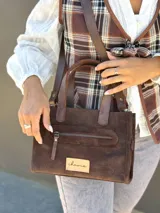 Cartera bandolera de cuero color marrón chocolate, con forro de tela y herrajes dorados. Tiene doble asa de mano y correa larga regulable y desmontable. Incluye bolsillo frontal con cierre y bolsillo interno.