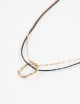Collar doble compuesto por una cadena fina metálica y un cordón negro, con un dije central de forma ovalada irregular en tono dorado.