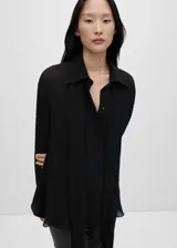 Camisa de tejido fluido y semitransparente, con cuello camisero, cierre frontal de botones, mangas largas con puños abotonados y lazo integrado en el cuello. Presenta un diseño recto con aberturas laterales en el bajo.