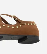 Zapatilla tipo Mary Jane color marrón con efecto gamuzado, tira fina con hebilla ajustable sobre el empeine y pequeñas tachuelas metálicas doradas. Tiene punta redonda y suela baja.