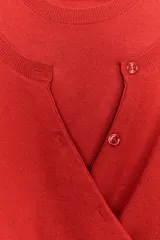 Chaqueta de punto color rojo con diseño de doble capa cruzada en el frente. Presenta cuello redondo, manga larga y cierre frontal con botones, con un acabado de tejido acanalado en el ruedo.