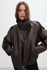 Cazadora tipo biker de cuero sintético en color marrón oscuro, con cuello subido, cierre frontal cruzado con botones forrados y cremallera oculta. Presenta mangas largas con trabillas en los hombros, bolsillos laterales de vivo y bajo con acabado elástico.