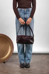 Cartera tipo baguette confeccionada en cuero de color marrón oscuro, con diseño alargado, cierre diagonal y correa de hombro.