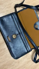 Cartera tipo bandolera de cuero metalizado en tono azulado, con diseño rectangular, solapa con cierre magnético y detalle de trenzado central. Incluye correa regulable y placa metálica con logo.