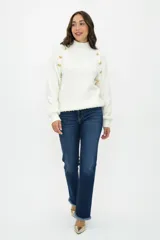 Buzo de tejido de punto texturizado en color blanco, con cuello alto y detalle de botones dorados decorativos en los hombros. Presenta mangas largas con puños acanalados y un corte recto.