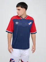 Remera de fútbol Umbro del Club Nacional de Football, de diseño tricolor con cuerpo azul marino, mangas y canesú rojos, y cuello en V con detalles en blanco y azul. Incluye el logo de Umbro en el pecho derecho y el escudo del club en el izquierdo.