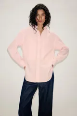 Camisa de corte oversize color rosa claro con diseño de rayas verticales sutiles. Presenta cuello solapa desmontable, cierre frontal con botones ocultos bajo solapa, mangas largas con puños y bajo asimétrico redondeado. Incluye dos bolsillos frontales amplios.