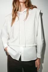 Camisa blanca de manga larga con un top de sudadera gris claro superpuesto, que incluye capucha ajustable con cordones. El bajo de la camisa tiene aberturas laterales.