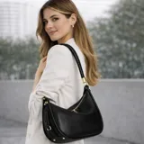 Cartera Guess modelo Danya de formato hobo, color negro con textura granulada. Presenta cierre superior, asa de hombro con detalle trenzado y logo metálico triangular en el frente.