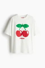 Remera de corte oversized en algodón, color blanco, con cuello redondo acanalado y diseño estampado de cerezas con el logo Pacha en el frente.