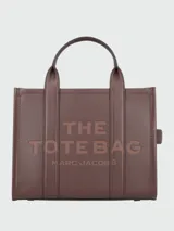 Cartera tipo tote de cuero granulado color tostado, con diseño estructurado y rectangular. Presenta doble asa superior fija y una correa larga ajustable y extraíble. En el frente, destaca el logo estampado con la inscripción 'THE TOTE BAG MARC JACOBS'.