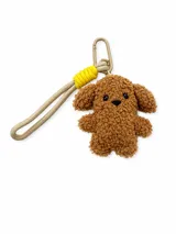 Llavero de peluche con forma de perrito color marrón, con correa de tela beige, detalles en amarillo brillante y gancho metálico.