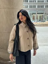 Chaqueta corta de gabardina en color beige, con cuello camisero, cierre frontal de botones y mangas largas con puños vueltos que revelan un forro a cuadros.