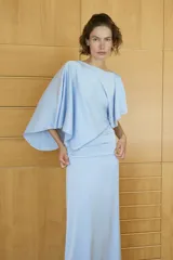 Vestido midi de tejido satinado color celeste, con cuello barco y diseño asimétrico con mangas tipo capa. Presenta un detalle drapeado en la cintura y abertura en la espalda.